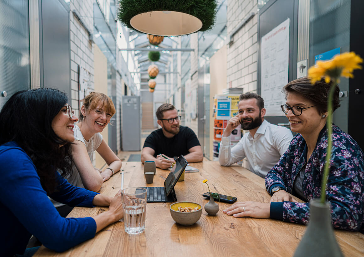 Eine Gruppe von fünf lächelnden Menschen sitzt an einem Holztisch in einem modernen Büro und diskutiert um einen Laptop.