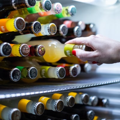 Eine Hand greift in ein Regal mit verschiedenen Flaschen, um eine Flasche mit orangefarbenem Etikett herauszunehmen.